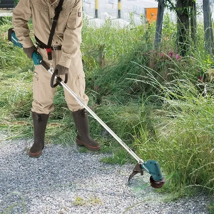 Roçadeira Makita DUR192LZ em uso, cortando mato alto ao lado de um caminho de cascalho